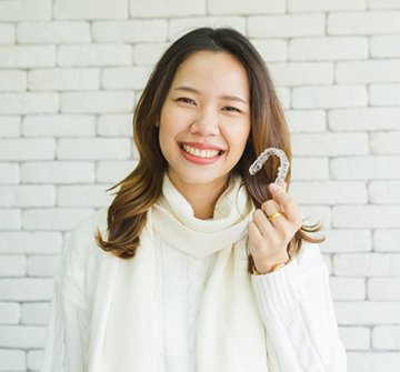 woman in white sweater holding up Invisalign aligner against white brick wall 